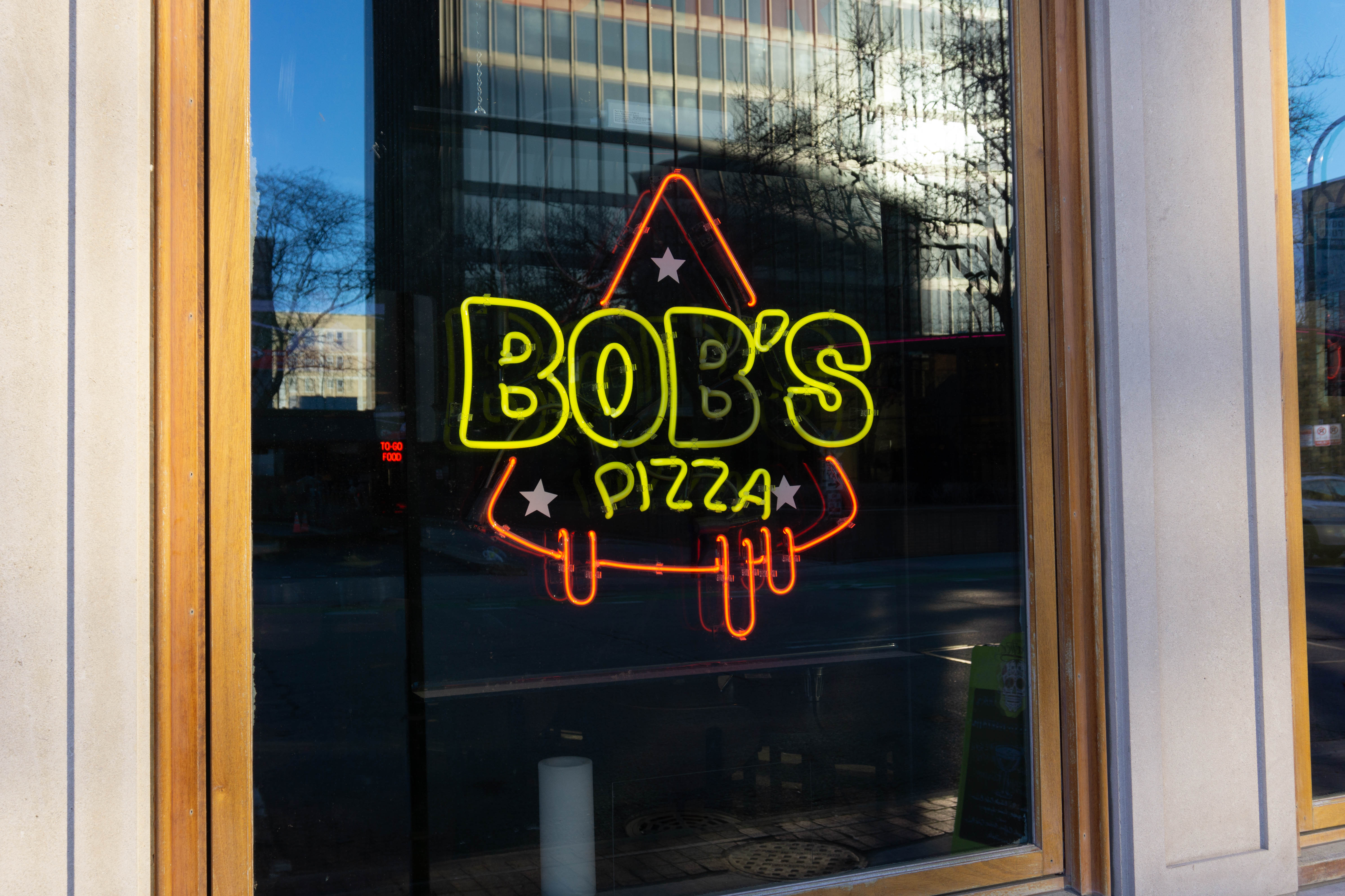 A neon sign in the shape of a pizza slice reads “Bob’s Pizza.”