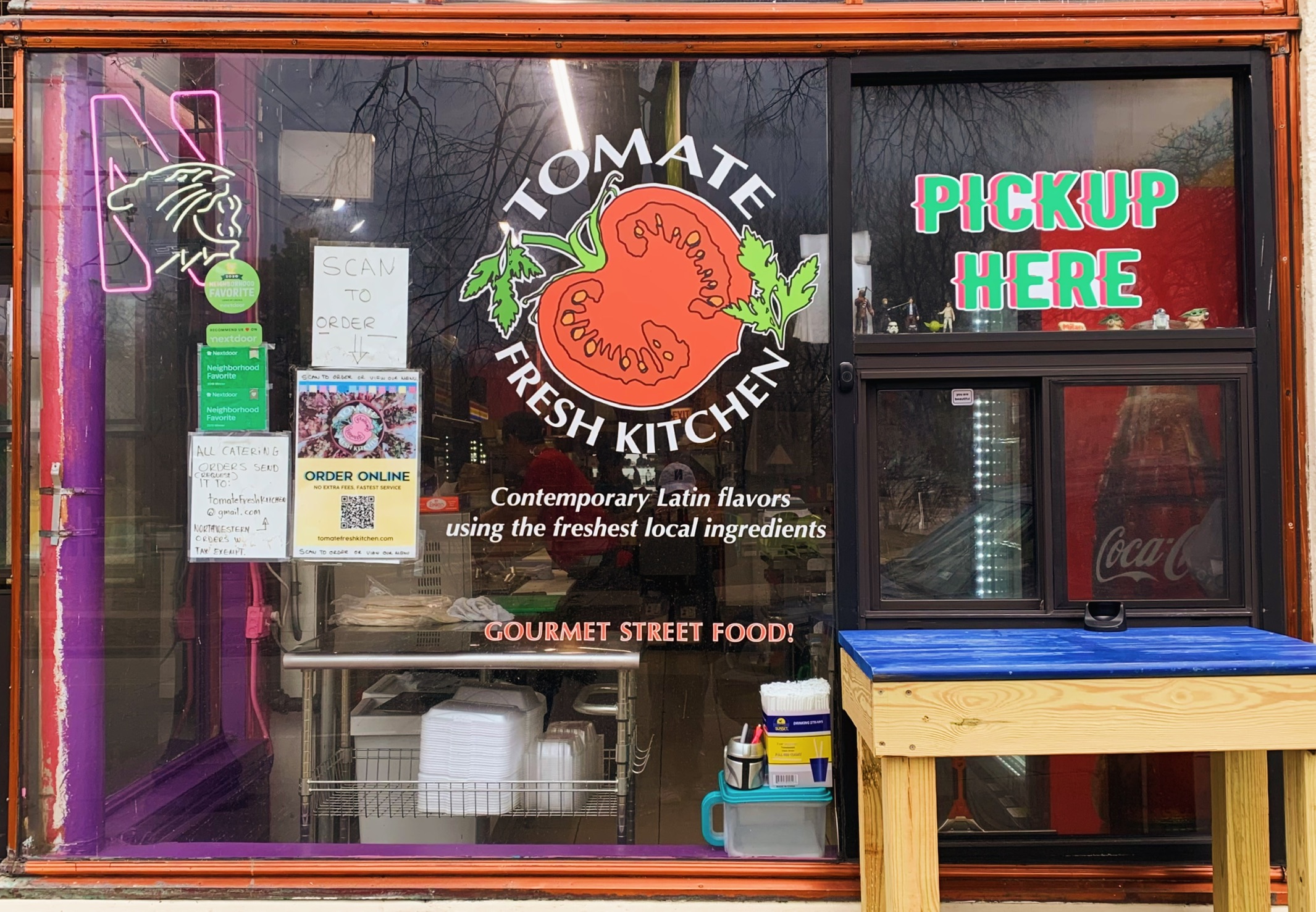 Tomate Fresh Kitchen storefront and pickup window. Signs and directions to order online are displayed next to the company's tomato logo. 