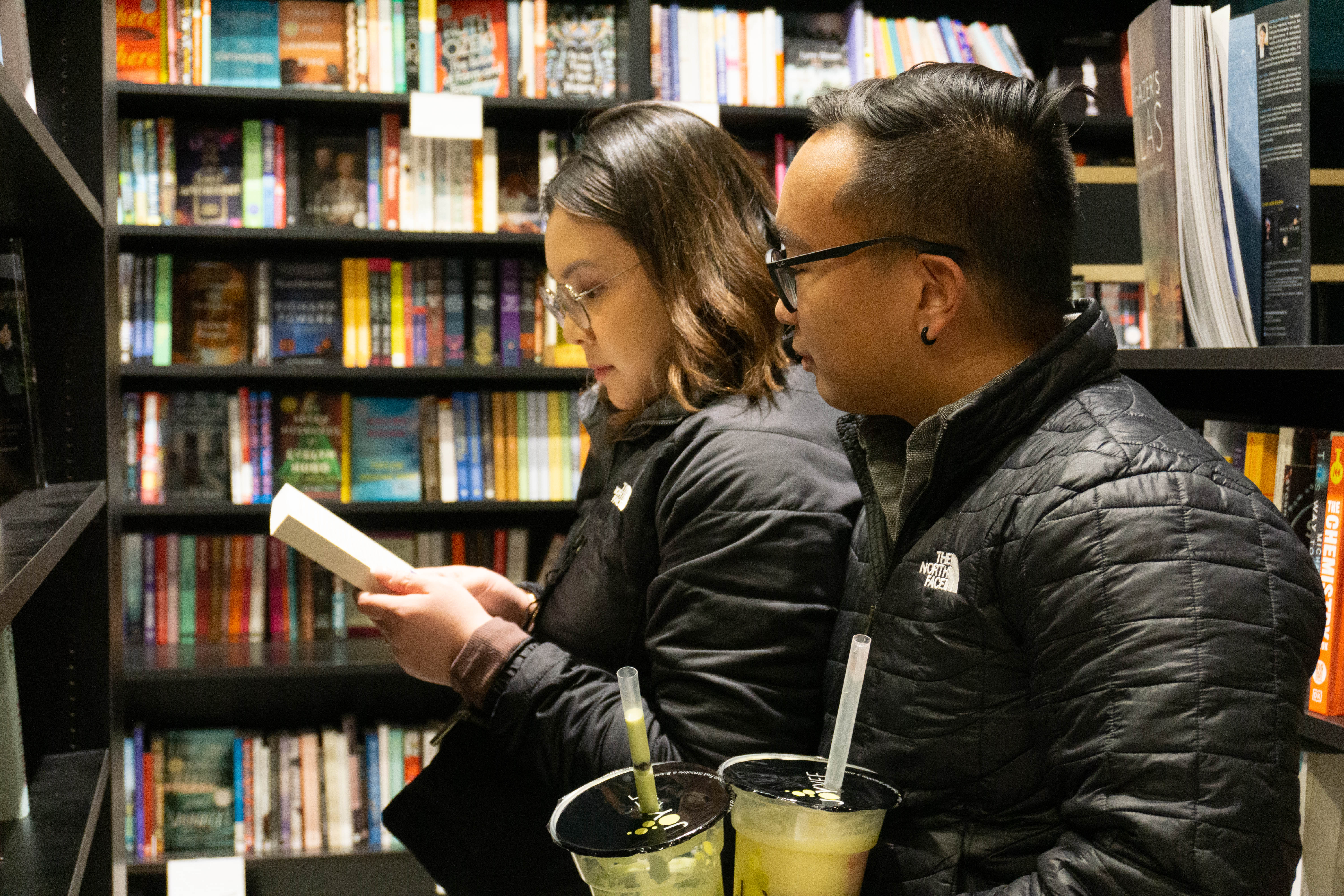 Two people in black jackets read a book while standing among bookshelves.