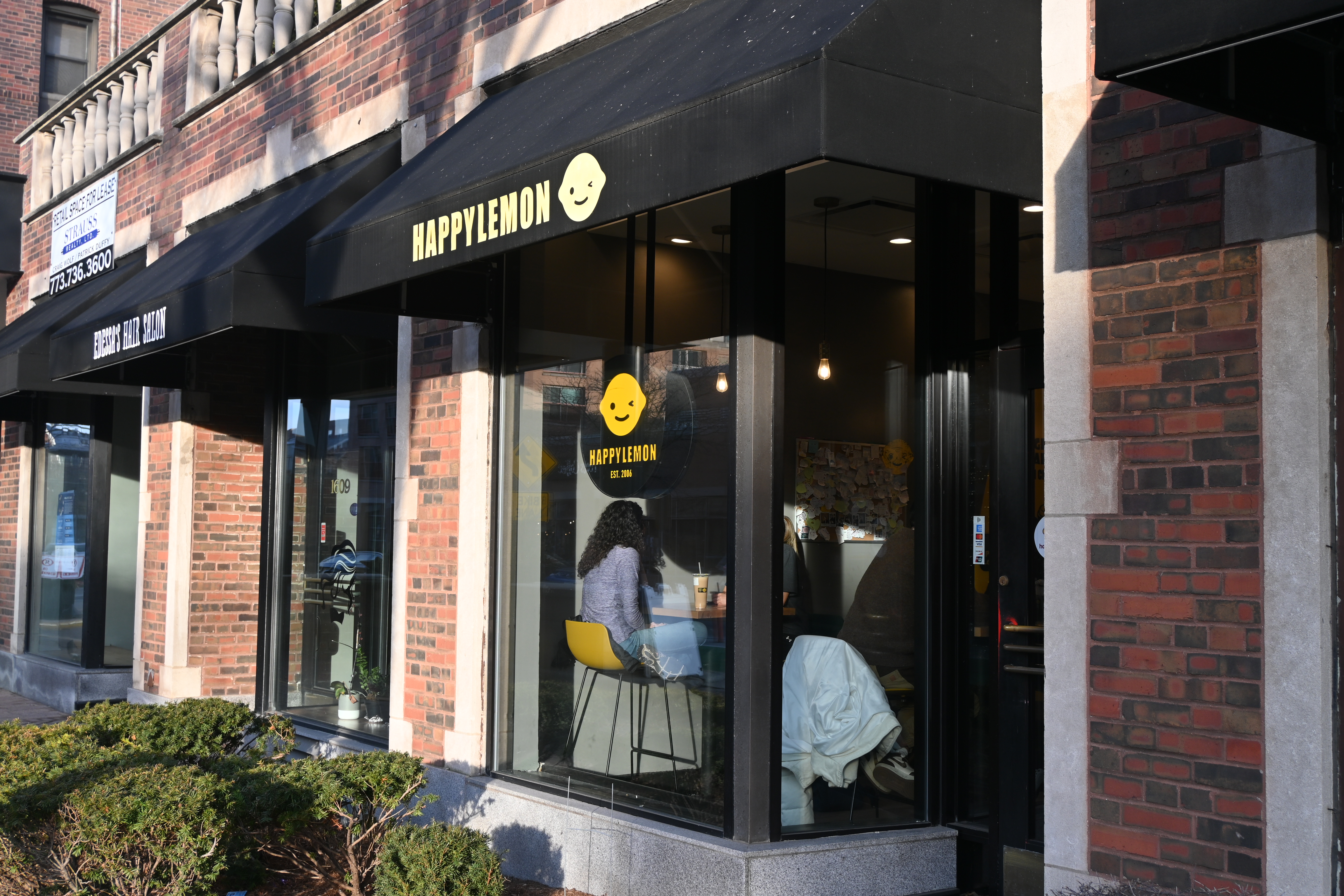 The front of a brick building with windows and a black sign that reads “Happy Lemon” in yellow lettering. 
							