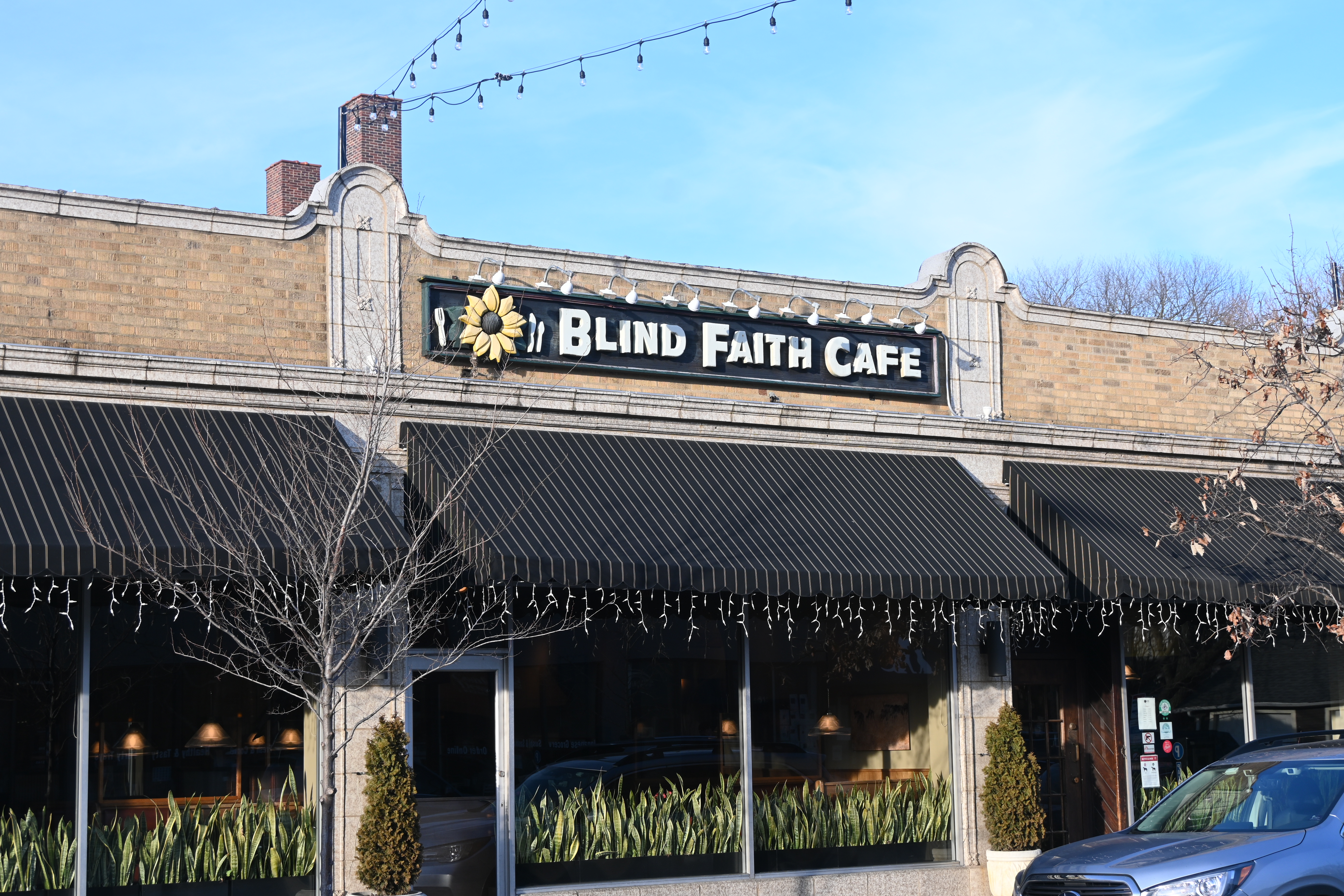 A black sign on a building reads “Blind Faith Cafe” in white letters next to a picture of a sunflower. A blue sky borders the building.