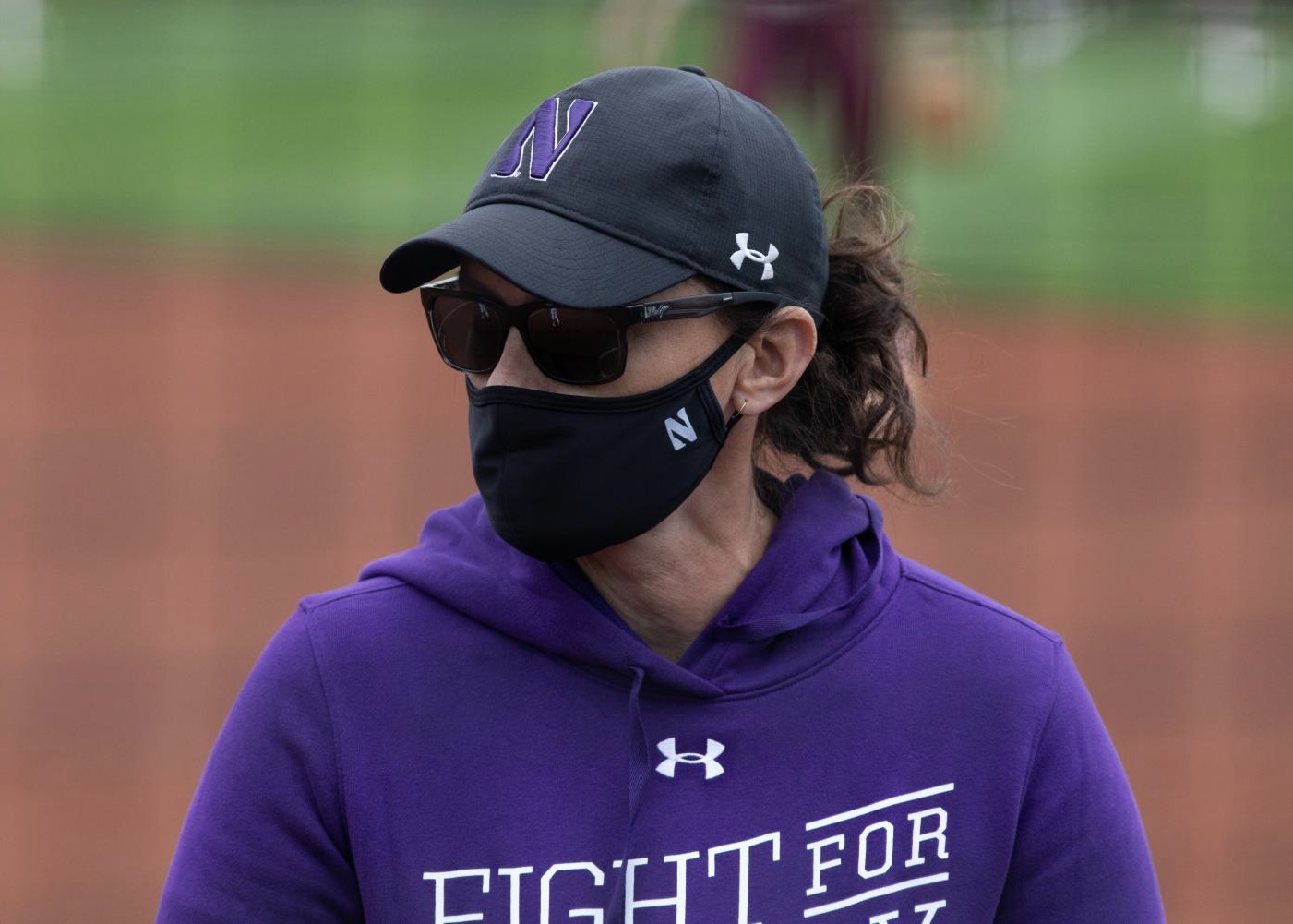 Woman in purple sweatshirts wearing mask looks at field.