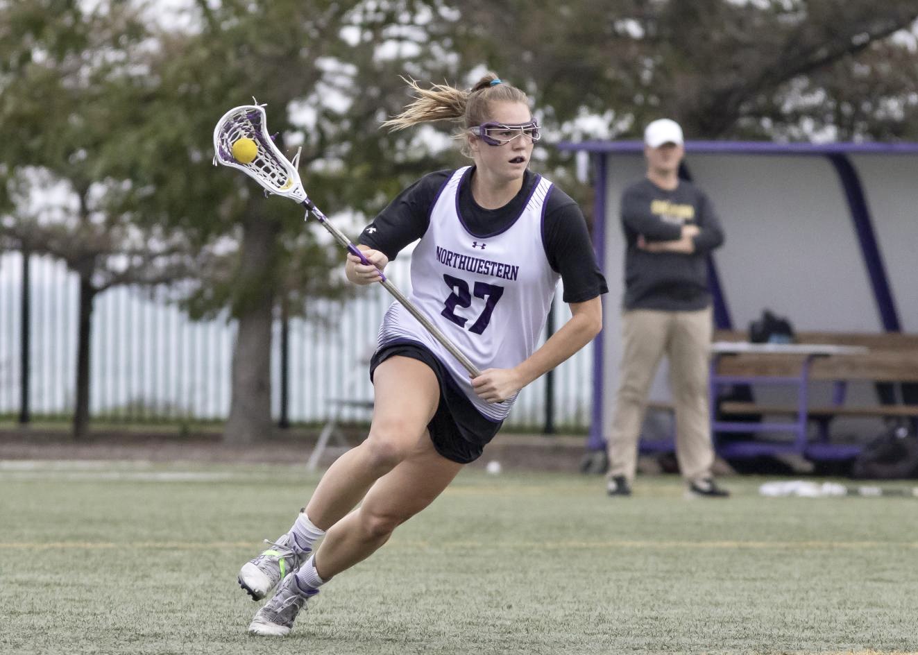 An athlete in a white jersey and black shorts runs while holding a lacrosse ball in a lacrosse stick.